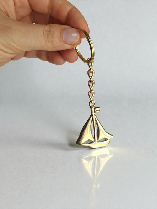 Gold sailboat keychain held by a hand on a light gray background