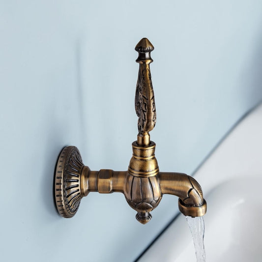 An ornate antique bronze wall-mounted faucet spout with a decorative handle and a circular backplate, shown with water flowing from the spout against a pale blue wall.