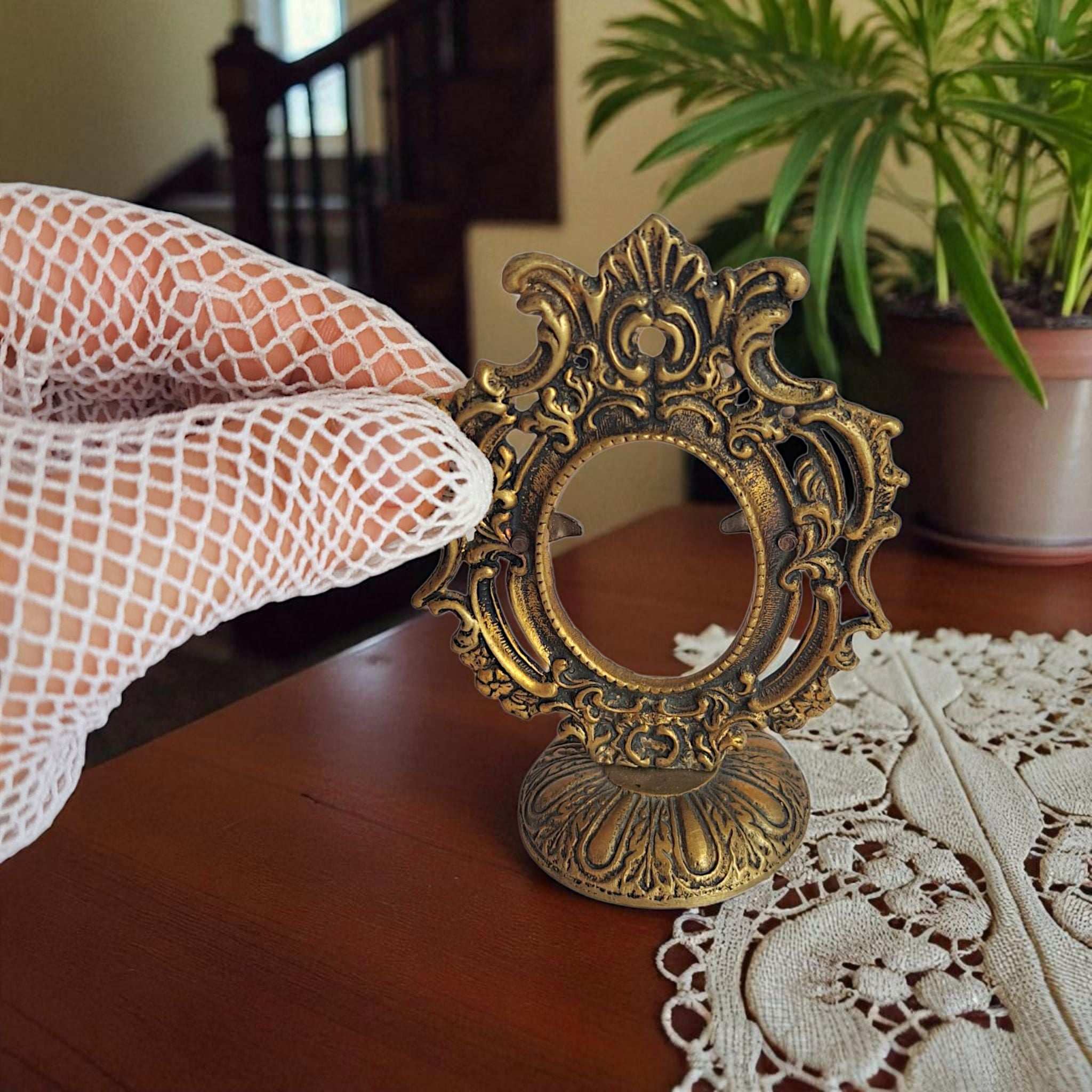 Decorative gold mirror held by a gloved hand on a lace tablecloth with a plant in the background.