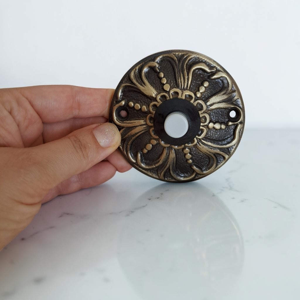 A hand holding a round, ornate antique bronze doorbell push button against a white marble surface and white background.