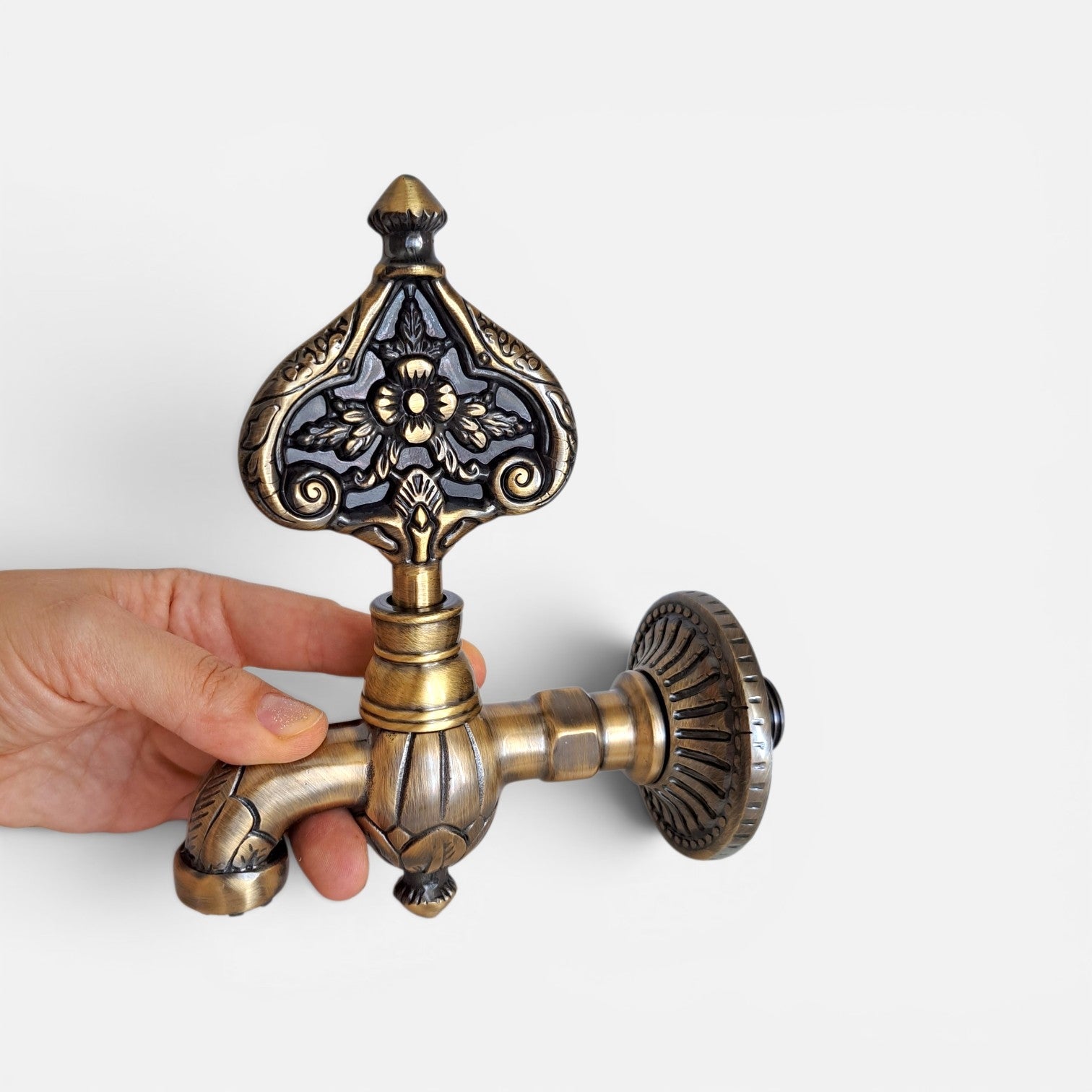 An ornate vintage-style brass faucet handle with a decorative floral pattern on the top knob and a ribbed round backplate, shown being held by a hand against a light gray background.