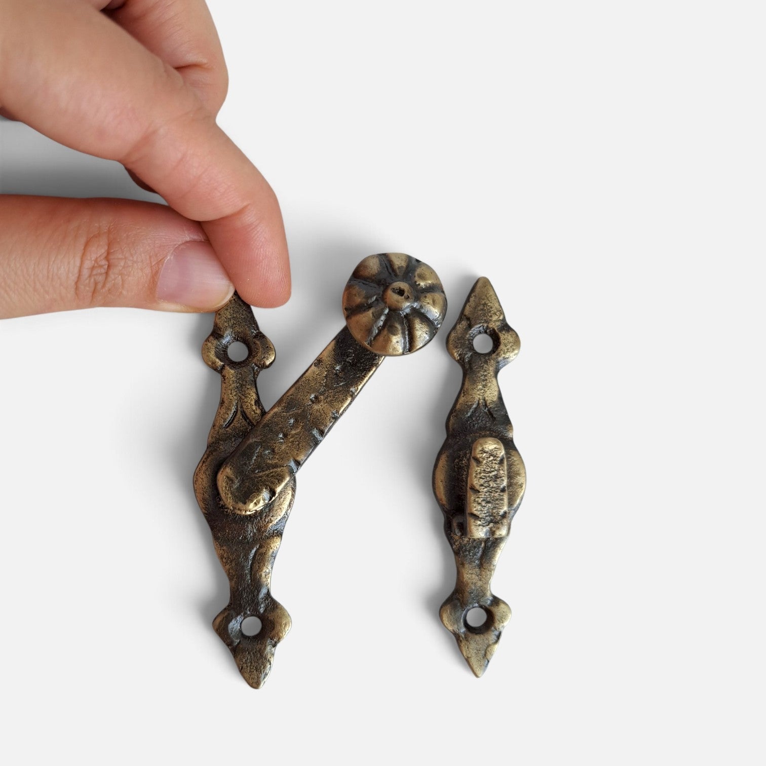 A close-up of a hand holding a pair of ornate antique-style metal hardware pieces against a plain light background.