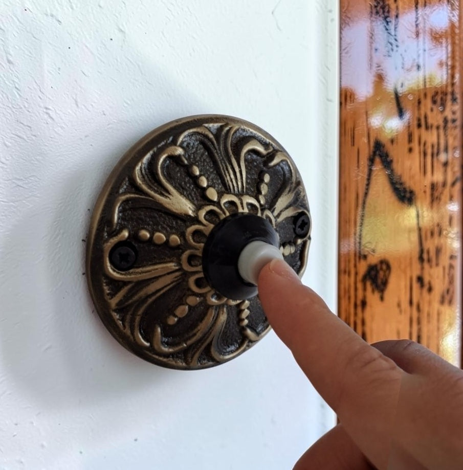 A close-up of a decorative round wall switch plate with an ornate embossed floral pattern in a dark bronze finish. A person's finger is pressing the center of the plate, which contains a black switch and a white cylindrical insert, mounted on a white textured wall.