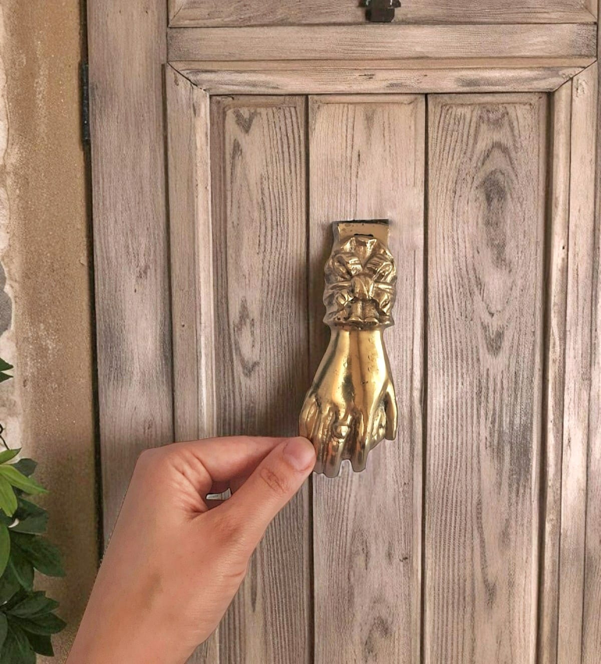 Hand holding a gold door knocker shaped like a hand on a wooden door.