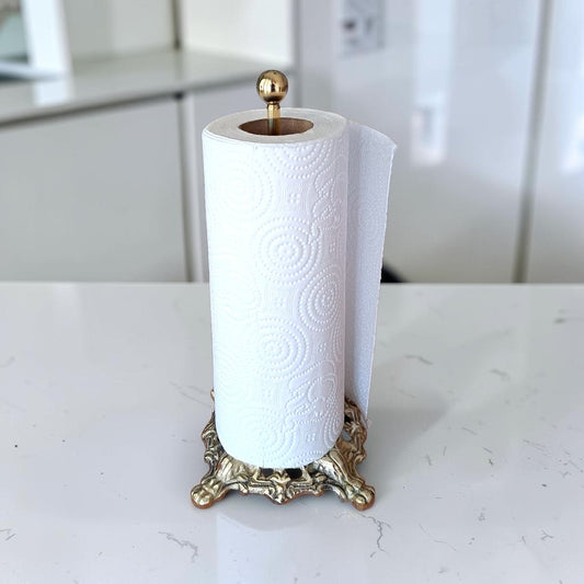 A roll of white patterned paper towels or kitchen paper on a decorative gold-toned stand, placed on a light marble countertop with a blurred kitchen background.
