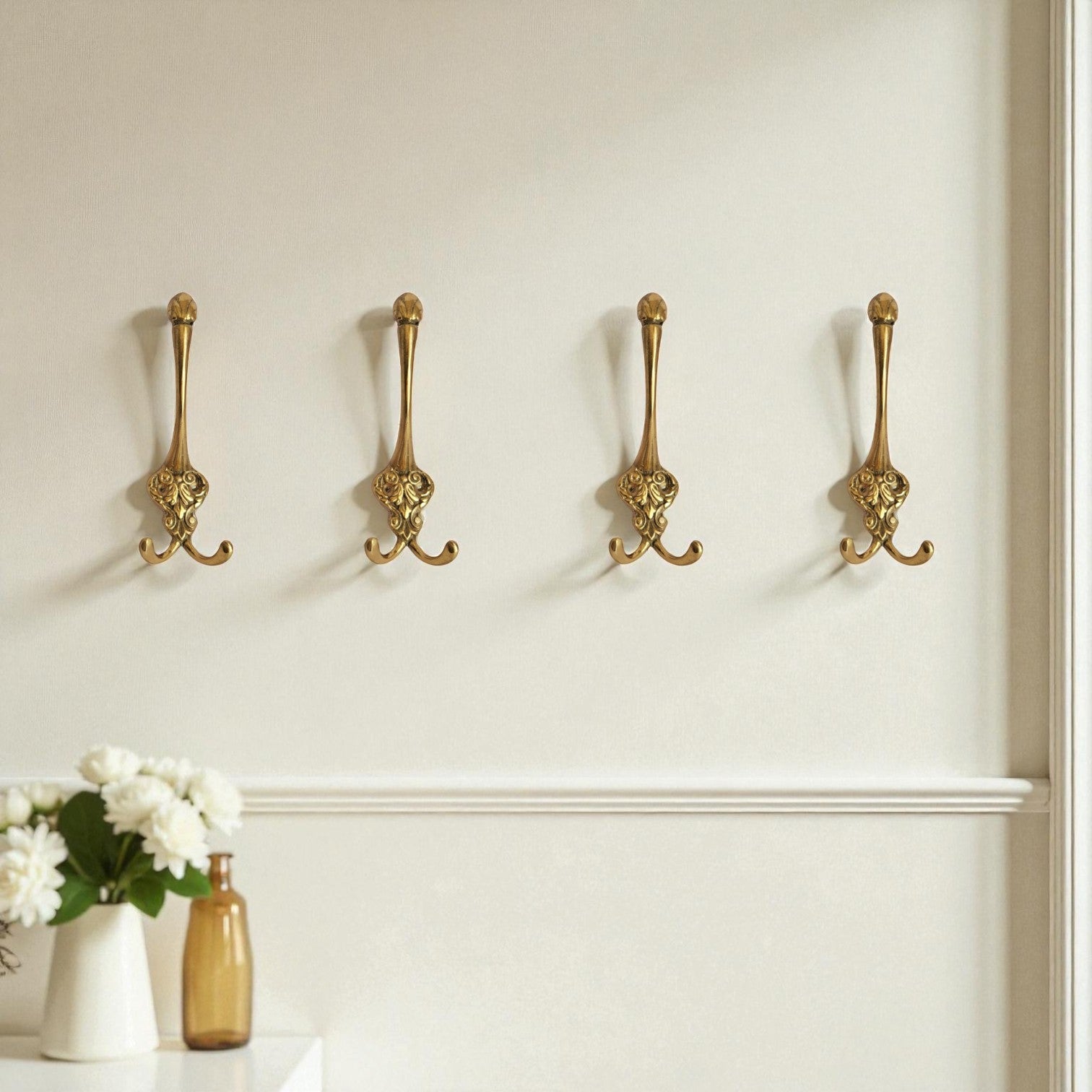 Four gold decorative hooks on a white wall above a shelf with a vase and flowers.