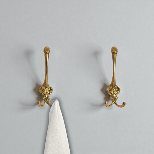 Two ornate gold wall hooks on a light gray wall with a white towel hanging from one of them.