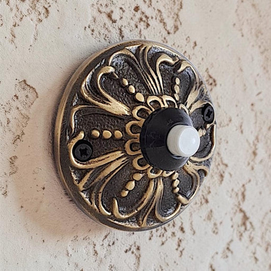 An ornate round metal doorbell push button with a decorative filigree design in antique bronze and gold tones, featuring a black central ring and a white button, mounted on a textured beige stucco wall.