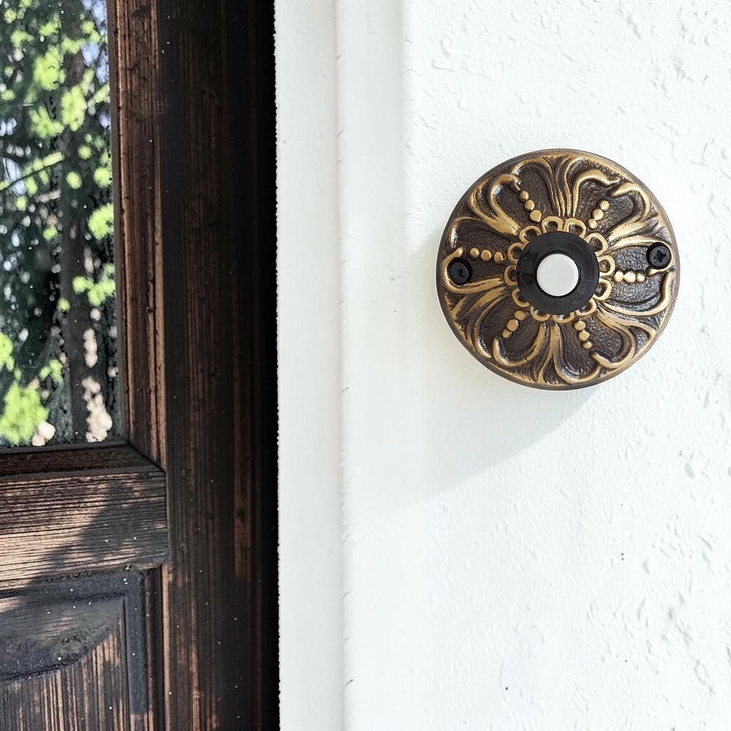 An ornate round doorbell push button with a bronze and gold floral pattern, mounted on a white textured wall next to a dark wooden door frame.
